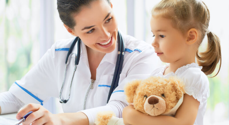 Photo of woman doctor sitting at her desk looking over medical results. Next to her is a young girl holding a teddy bear. The doctor is looking at the girl and talking about the results of peanut immunotherapy.