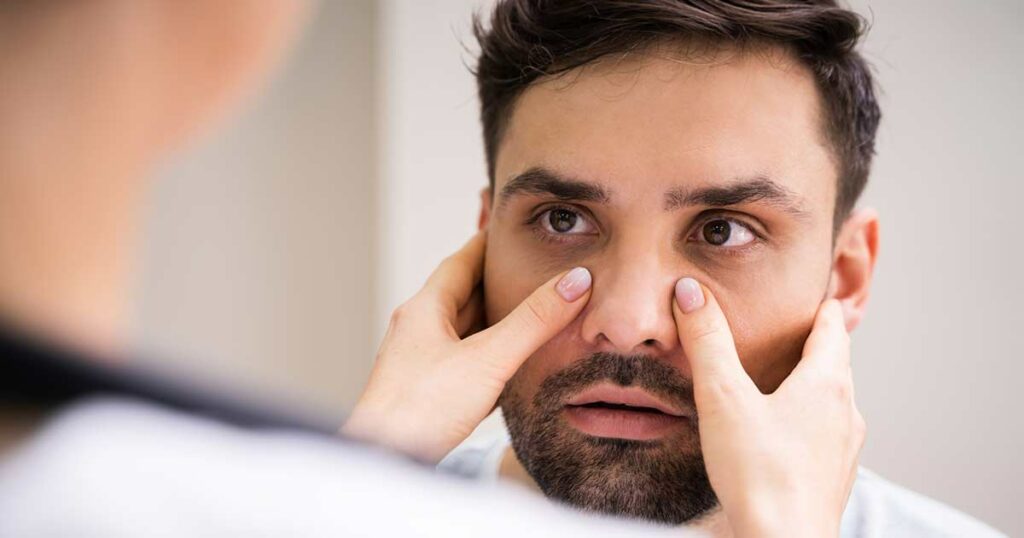 Face of a man looking at the doctor. Doctor is seen from the back with her hands on his face to feel his sinus inflammation.