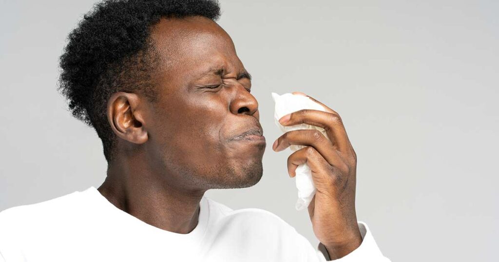 Man with face in visible discomfort is getting ready to hold a tissue to his nose because his sinuses are hurting.
