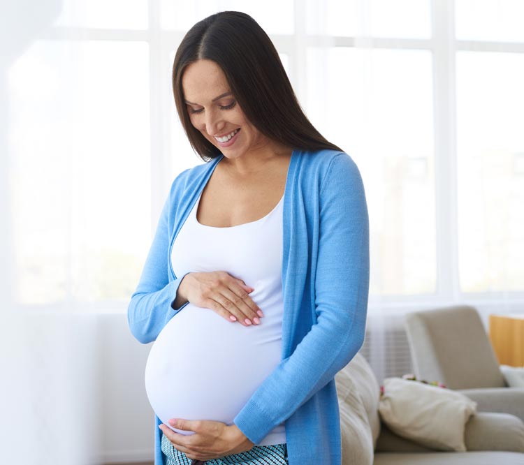 Woman in her living room holding her pregnant belly, looking down and smiling.