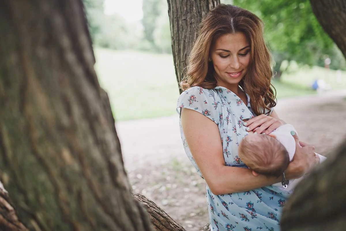 Latina woman discreetly breastfeeding her baby in the park, under a tree. She is smiling and looking at the baby.